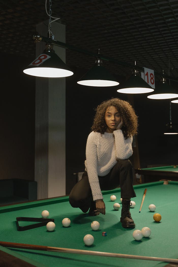 Woman with curly hair posing on a billiard table in dimly lit room.